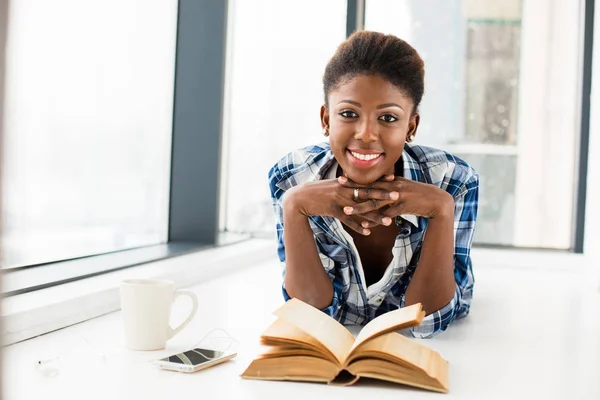 Mujer negra leyendo un libro al lado de una gran ventana con labio natural — Foto de Stock