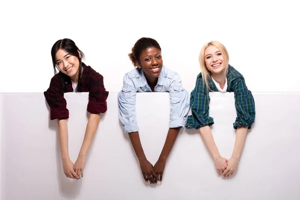 Três diferentes meninas étnicas apoiando-se no painel branco — Fotografia de Stock