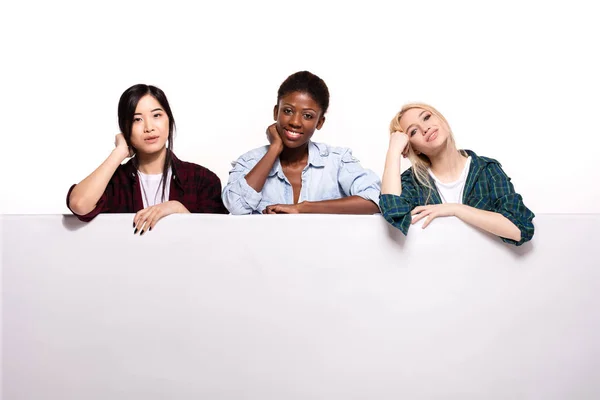 Três diferentes meninas étnicas apoiando-se no painel branco — Fotografia de Stock