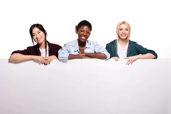 Três diferentes meninas étnicas apoiando-se no painel branco — Fotografia de Stock