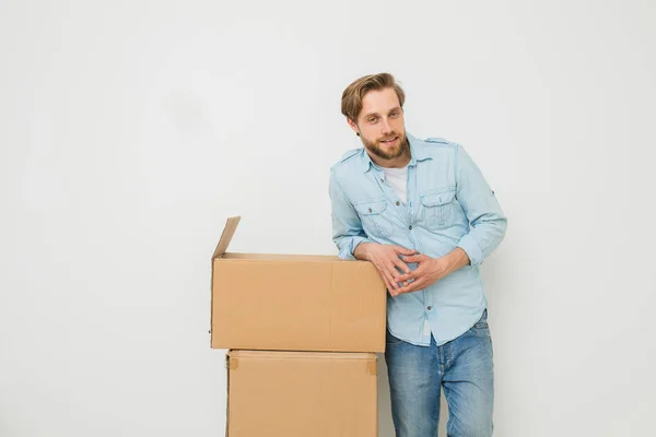 blonde man with beard standing relaxed next to his things packed in cardboard boxes, whaiting for someone to carry them, on white background