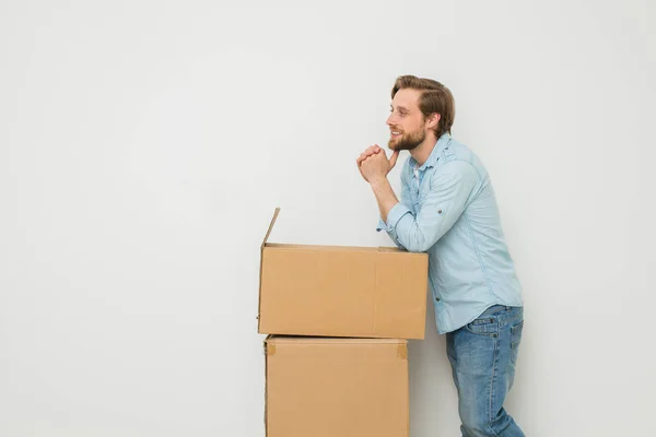 blonde man with beard standing relaxed next to his things packed in cardboard boxes, whaiting for someone to carry them, on white background, and looking to side with smile