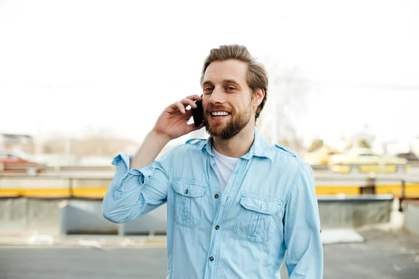 どこか外に笑みを浮かべて この会話を楽しみながら 携帯電話で話している会話を青いシャツを着てカジュアルな若い男 — ストック写真