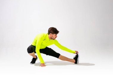 Önce esneme hareketleri yap. Spor giyim eğitimi alan genç bir adam beyaz arka planda kaslarını esnetiyor.