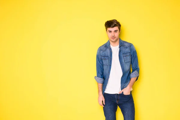 黄色の背景に立っているジーンズの服を着たクールなハンサムな若い男性 — ストック写真