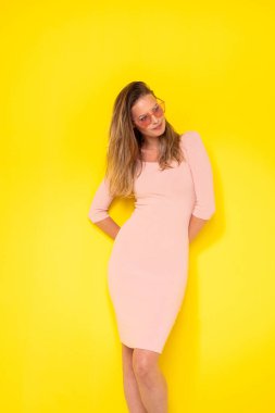 Genç ve modern kadın, seksi elbiseli, sarı arka planda sevinçle poz veren, pembe güneş gözlükleriyle yaz atmosferine ilham veren