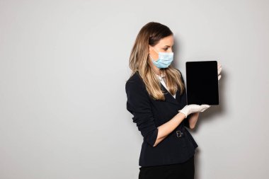 Resmi ofis kıyafetli, koruma maskeli ve eldivenli bir kadın kameraya tablet sunuyor.