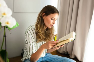 Parlak bir pencerenin yanında kitap okuyan, salgın sırasında dinlenen ve dinlenen bir kadın. Sağlıklı zihin için ev aktivitesi