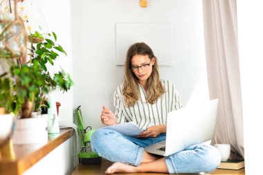 Evden ya da internetten çalışmak, evin içinde dizüstü bilgisayarda çalışan bir kadınla, parlak bir pencerenin yanında ve ev bitkisinin yanında çalışmak.