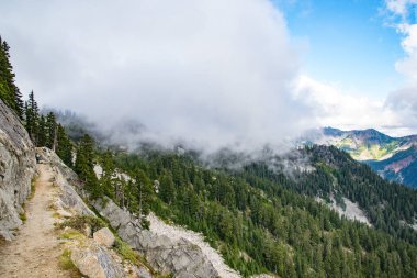 Dağları yeşil ormanlarla kaplayan dev bir bulut ve kenarlarda dar yürüyüş yolları.