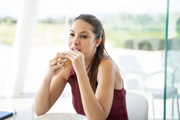 坐在明亮的窗前同事的办公室里 一个带着尾巴的年轻女人正在吃三明治 — 图库照片