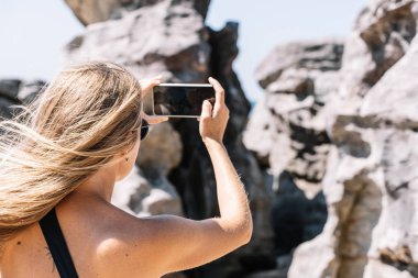 Sarışın kız sahilde kayalıklarda cep telefonuyla fotoğraf çekiyor.