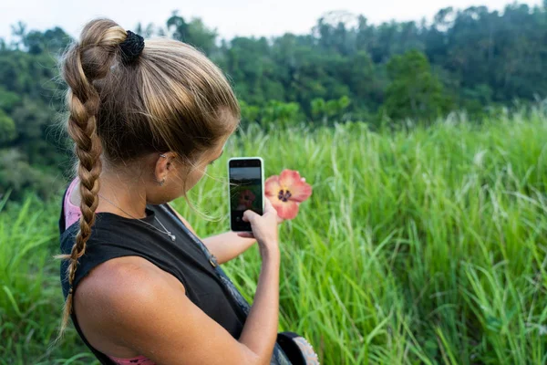 Blonde woman with a braid taking a picture with her mobile phone of a flower she is holding in her hand in the middle of a green field