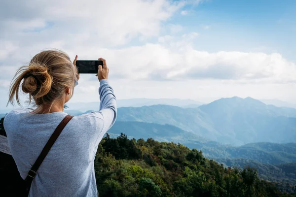 Blonde girl taking pictures with the mobile of the horizon views in the mountains with the cloudy sky