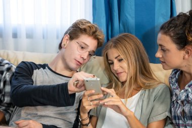 Gayri resmi olarak giyinmiş bir grup genç, yemek odasındaki bir kanepede oturup birlikte cep telefonuna bakıyorlar.