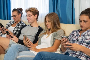 Gayri resmi olarak giyinmiş bir grup genç, yemek odasındaki bir kanepede oturup birlikte cep telefonuna bakıyorlar.