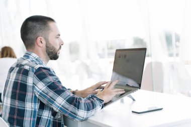 Dizüstü bilgisayarın önünde çalışan sakallı genç bir adam. Bir tarafında cep telefonu olan parlak ışıklandırılmış bir iş arkadaşı.