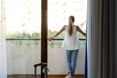 Beyaz perdeli bir otel odasının korkuluklarına yaslanmış genç sarışın kız.