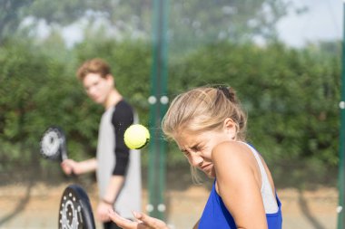 Top sarışın bir kızın alnına raketle vuruyor açık hava raket kortunda genç bir çocuğun önünde.