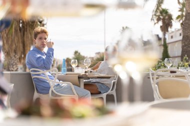 Gözlüklü ve piercingli genç bir adam yanında sarışın bir kızla birlikte bir restoranın teras masasında sigara içiyor.