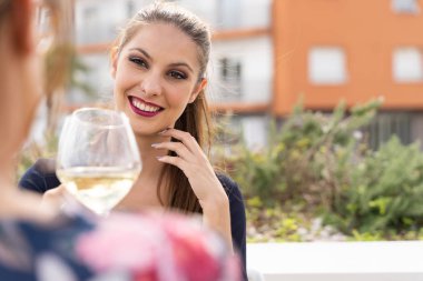 Restoran terasında başka bir kızla kadeh kaldırırken gülümseyen bir kadın. Etrafı binalar ve çalılarla çevrili.