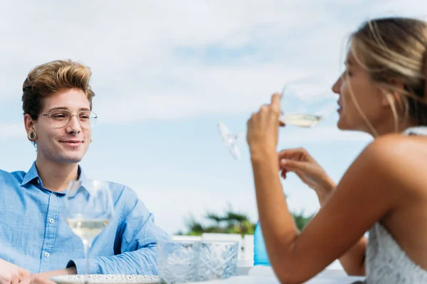 海に面したレストランのテラスでガラスから水を飲んでいる金髪の女の子を見るピアスとメガネを持つ若い男 — ストック写真