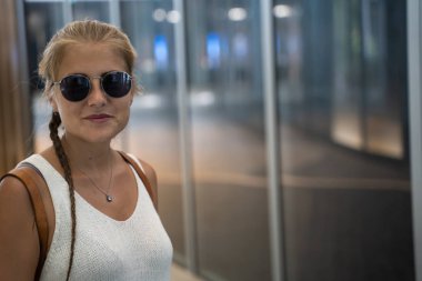 Sarışın, güneş gözlüklü, örgülü ve beyaz atletli bir kız Singapur 'da camdan bir cephesi olan modern bir binanın önünde duruyor.