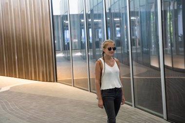 Sarışın, güneş gözlüklü, örgülü ve beyaz atletli bir kız Singapur 'da camdan yüzüyle modern bir binanın önünde yürüyor. Seyahat kavramı