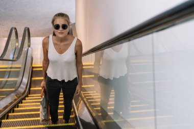 Singapur 'da modern bir binada yürüyen merdivene tırmanan güneş gözlüklü ve el çantalı sarışın bir kız.