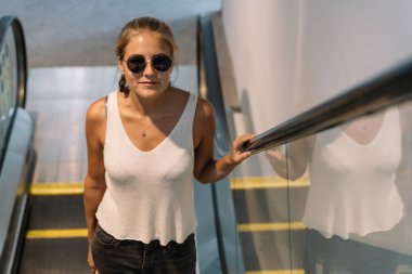 Singapur 'da modern bir binada yürüyen merdivene tırmanan güneş gözlüklü sarışın kız.