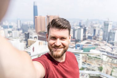 Yaz kıyafetleri içinde sakallı bir adam Malezya 'da Kuala Lumpur' da arka planda bir şehirle dışarıda selfie çekiyor.