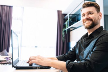 Genç sakallı bir adam dizüstü bilgisayarla uzaktan çalışıyor modern bir odada ahşap bir masada oturuyor. Dijital Göçebe kavramı