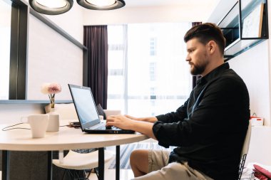 Modern bir odada ahşap bir masada dizüstü bilgisayarla uzaktan çalışan sakallı bir adam. Dijital Göçebe kavramı