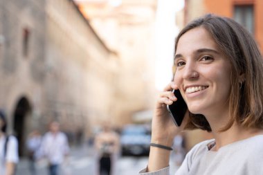 Yarım yelesi ve delinmiş burnu olan, gülümseyen ve sokakta cep telefonuyla konuşan genç bir kadın.