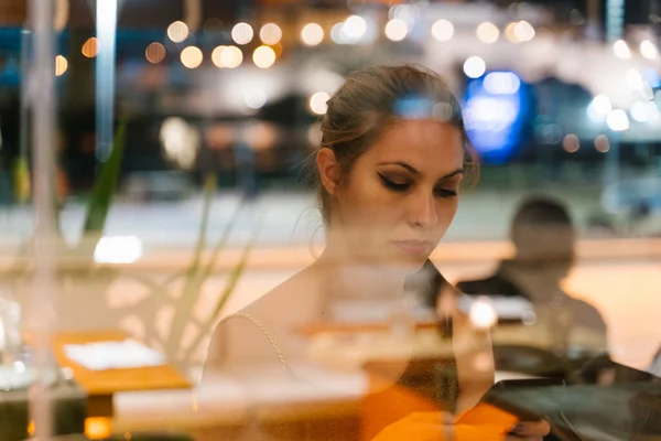 Young woman sitting inside a restaurant using her mobile phone with serious expression and the street lights reflected in the glass at night