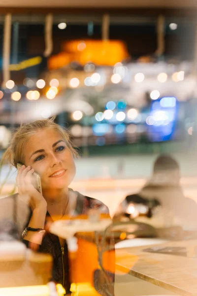 金发女人坐在餐馆里 带着严肃的表情用她的手机与人交谈 街上的灯光反射在夜晚的玻璃上 — 图库照片