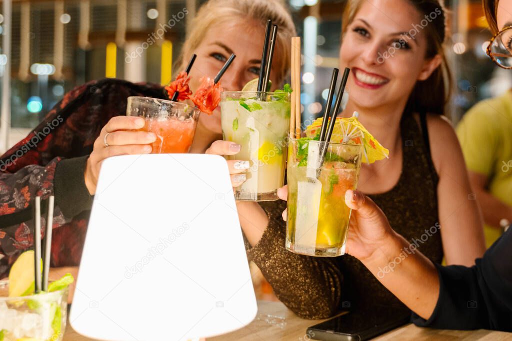 Selective focus on the glasses of cocktail of three woman making a selfie while sitting on a terrace drinking cocktails at night