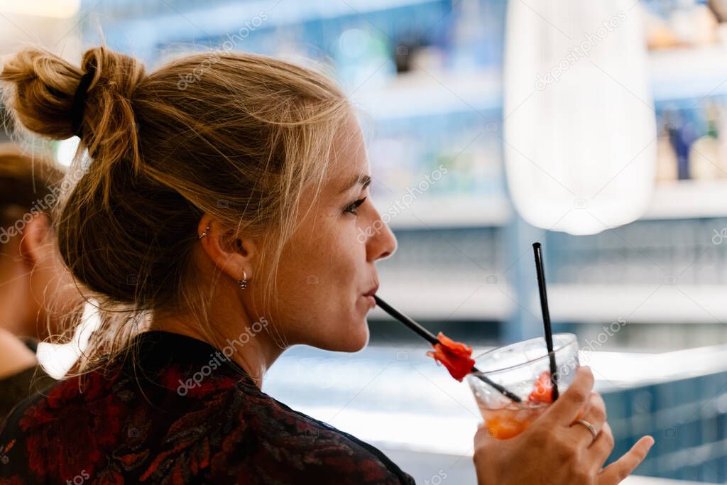 Profile of a blonde woman on her back sitting at a bar and sucking a straw of a cocktail