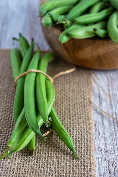 Close-up of green beans on burlap tied with rope, with more beans in wooden bowl, out of focus, vertically