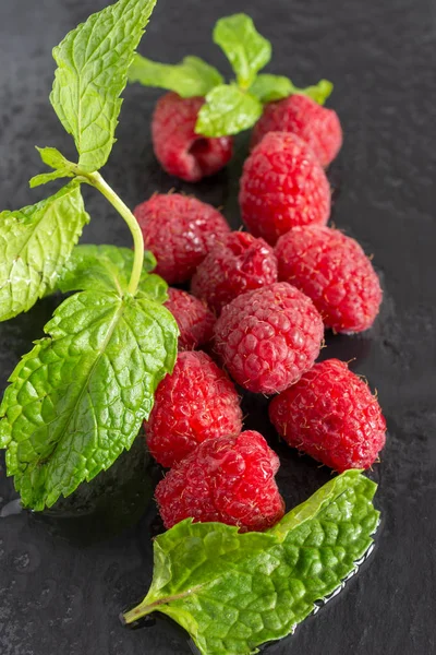 Top view of wet raspberries and mint leaves out of focus on black slate in vertical