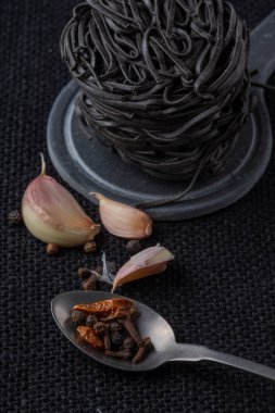 Sarımsak yakın plan, baharatlı metal kaşık ve siyah tagliatelle, siyah çuval bezi arka plan, dikey