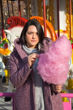 Orta boy esmer bir kadın, pembe pamuk şekerle poz veriyor, atlıkarıncanın önünde, gün batımı ışığında, dikey