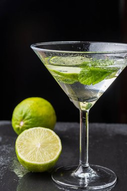 Nane yapraklı ve limon dilimli kokteyl bardağını yakın plan, siyah zemin üzerinde bir buçuk misket limonu ve dikey odaklı