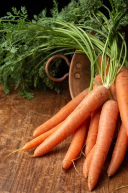 Yeşil yapraklı bir grup havuç yakın plan, bakır kâse, eskimiş ahşap masa, siyah arka plan, dikey