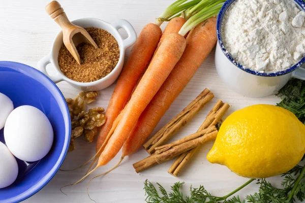 Aerial view of ingredients for sponge cake with carrots, eggs, carrots, flour, lemon, brown sugar, on white wooden table, horizontal