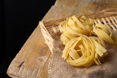 Çuval bezinde, sepette, kırsal masada ve siyah arkaplanda, yatay olarak çiğ tagliatelle görünümü