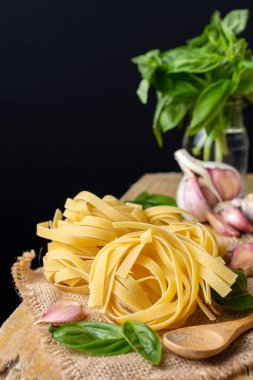 Çiğ tagliatelle yuvalarını tahta kaşık, fesleğen ve sarımsak ile çuval bezi üzerine yakın plan, kırsal masa ve siyah arka plan üzerine seçici odak,