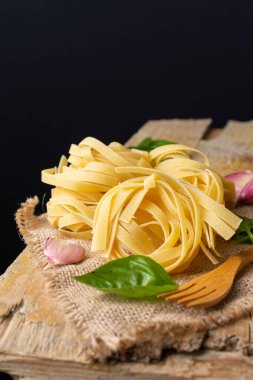 Çiğ tagliatelle yuvalarının üst görüntüsü ahşap çatal, fesleğen ve sarımsak, seçici odak, kırsal masa ve siyah arka plan, dikey