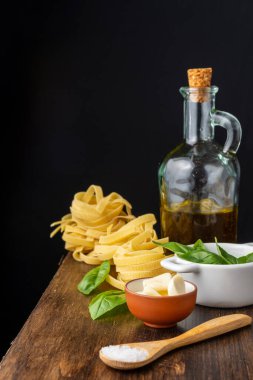 Tahta kaşık, tuz, sarımsak, fesleğen, zeytinyağı ve çiğ tagliatelle şişesi, ahşap masa, siyah arka plan, dikey
