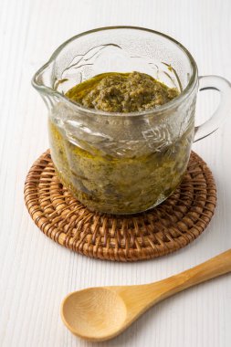 Cam kavanozun üstünde pesto sosu, tahta kaşık, beyaz tahta masa, dikey